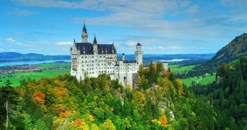 Castillo de torres puntiagudas en lo alto de una colina rodeado de bosques y montañas